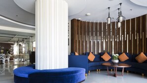 A modern lobby with a curved blue seating area, a round wooden coffee table, and a vertical wooden panel wall with hanging pendant lights.