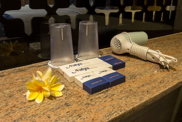 Un plan de travail en granit orné d’une fleur, deux verres transparents, un sèche-cheveux blanc et des distributeurs de savon à shampoing de la marque Matya Hotel.
