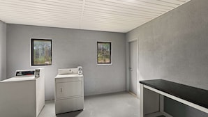 A laundry room with two washing machines, a dryer, and a countertop. There are two windows with views of trees. - SureStay by Best Western Gulfport (Gulfport)