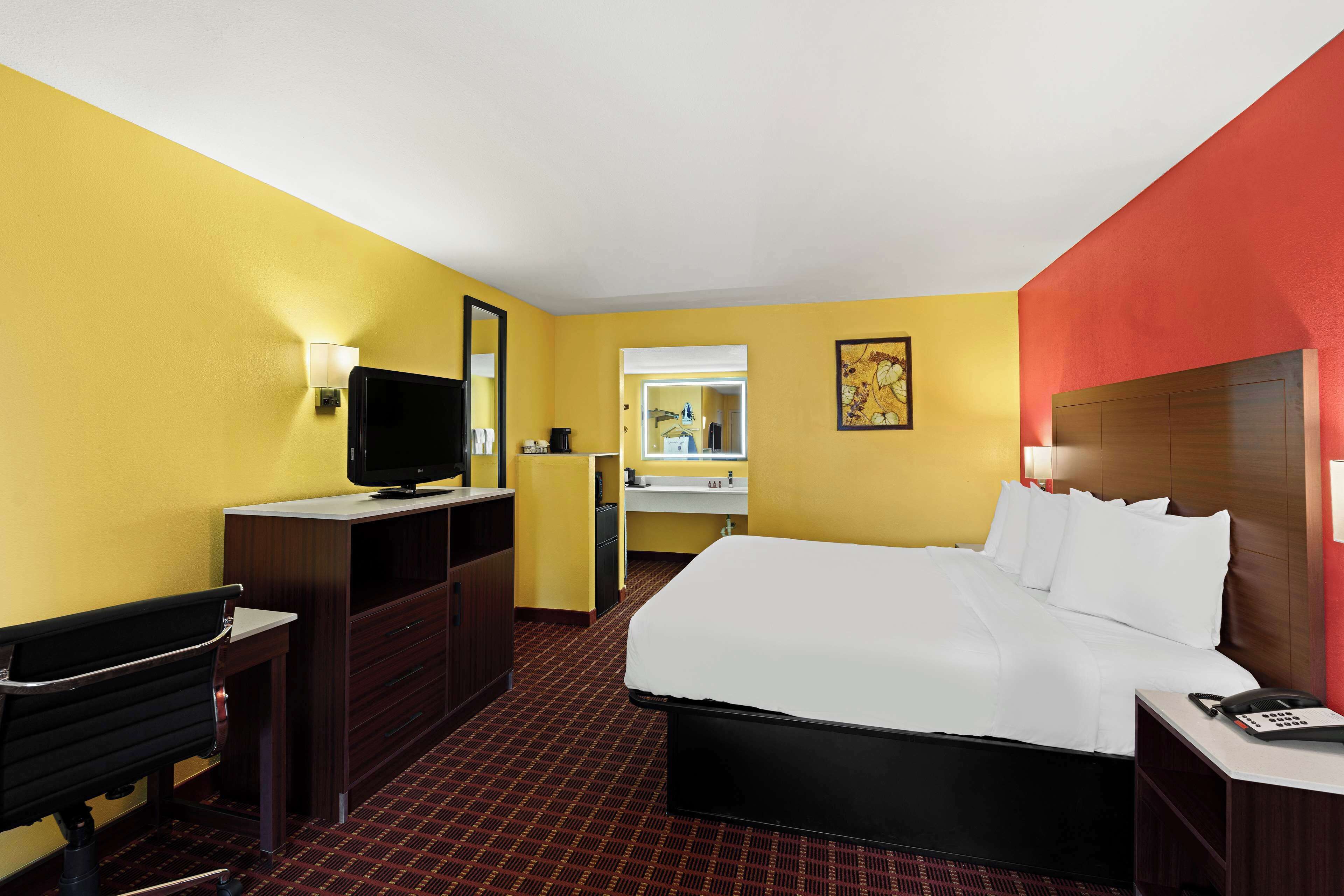 Ein Hotelzimmer mit einem Bett, einem Schreibtisch, einem Stuhl, einem Fernseher, einem Spiegel und einem durchgegangenen Tür ersichtlichen Badezimmer.