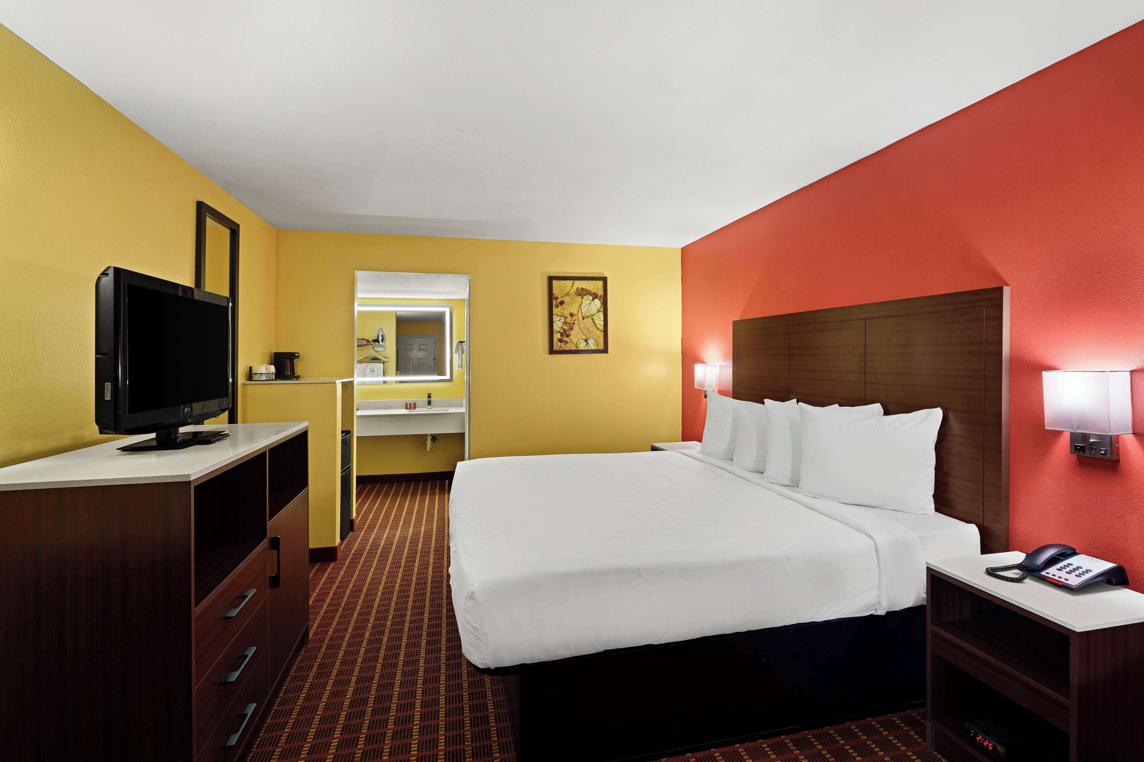 A hotel room with a bed, a TV, a desk, a mirror, and a bathroom visible through an open door.