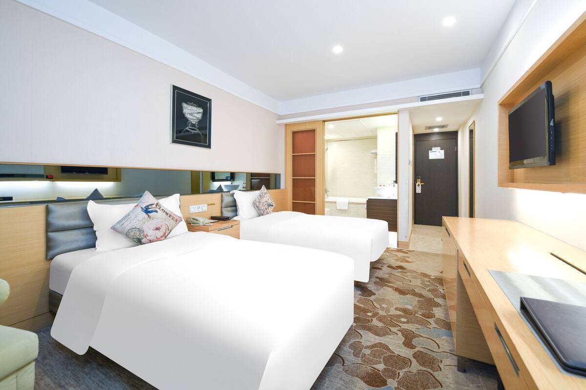 Habitación de hotel con dos camas, un escritorio, un televisor y un baño visible a través de una puerta abierta.