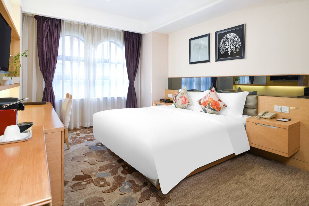 Habitación de hotel con una cama grande, una mesita de noche con lámpara, un pequeño escritorio con teléfono y una ventana con cortinas.