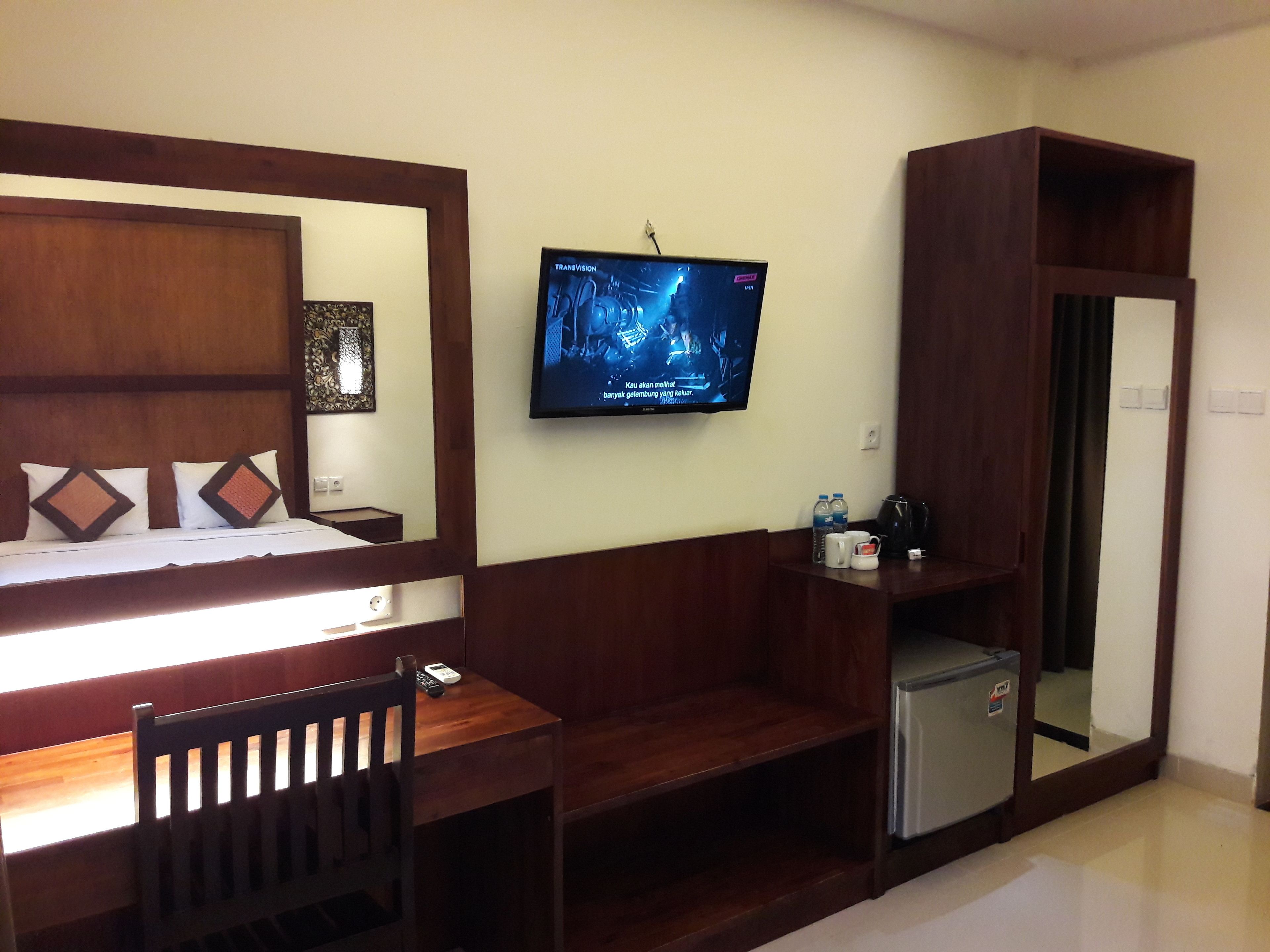A hotel room with a bed, a desk, a chair, a TV mounted on the wall, a small refrigerator, and a mirror.
