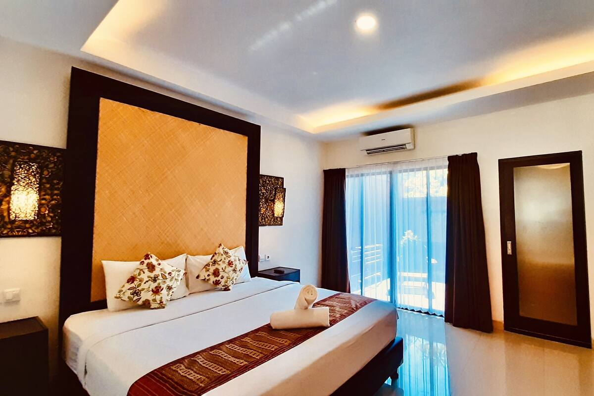 A hotel room with a large bed, a nightstand, a wall-mounted air conditioner, and a sliding glass door leading to a balcony.