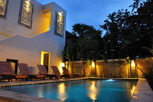 Una piscina con sillones de descanso e iluminación decorativa, rodeada por un edificio moderno y plantas tropicales.