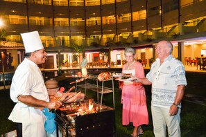 Un chef preparando comida a la parrilla al aire libre, con invitados cerca.