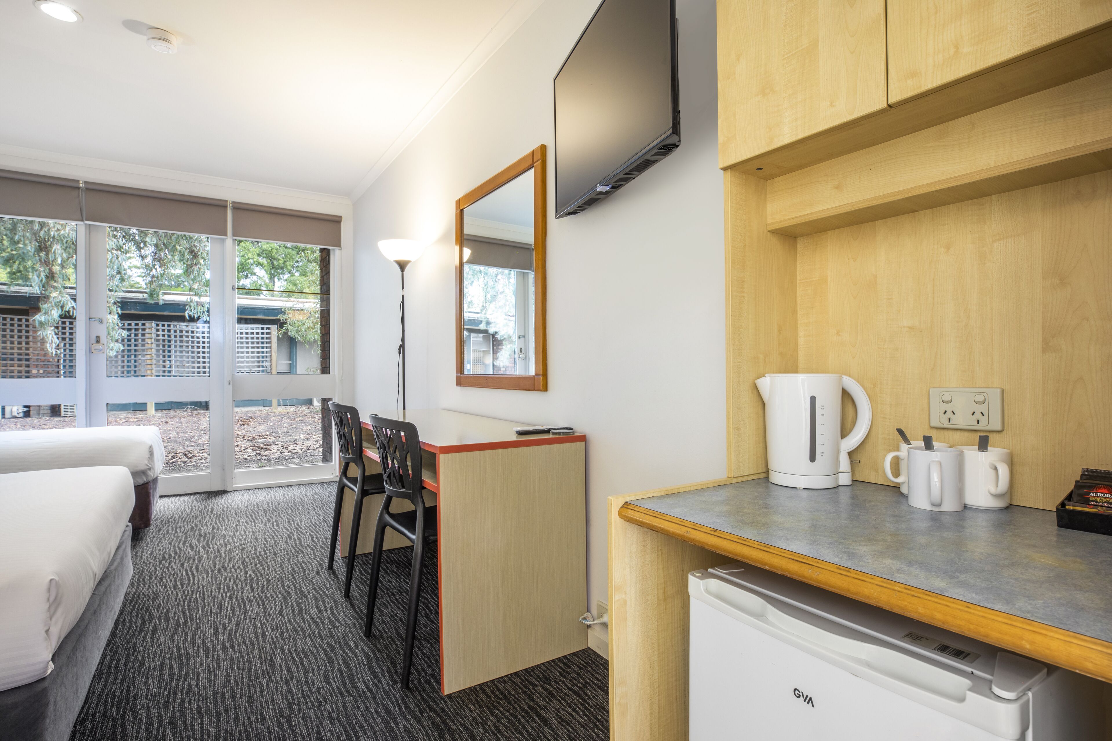 A hotel room with a bed, a desk with a chair, a kitchenette with a coffee maker and mugs, a TV mounted on the wall, and a large window with a view of the outdoors.