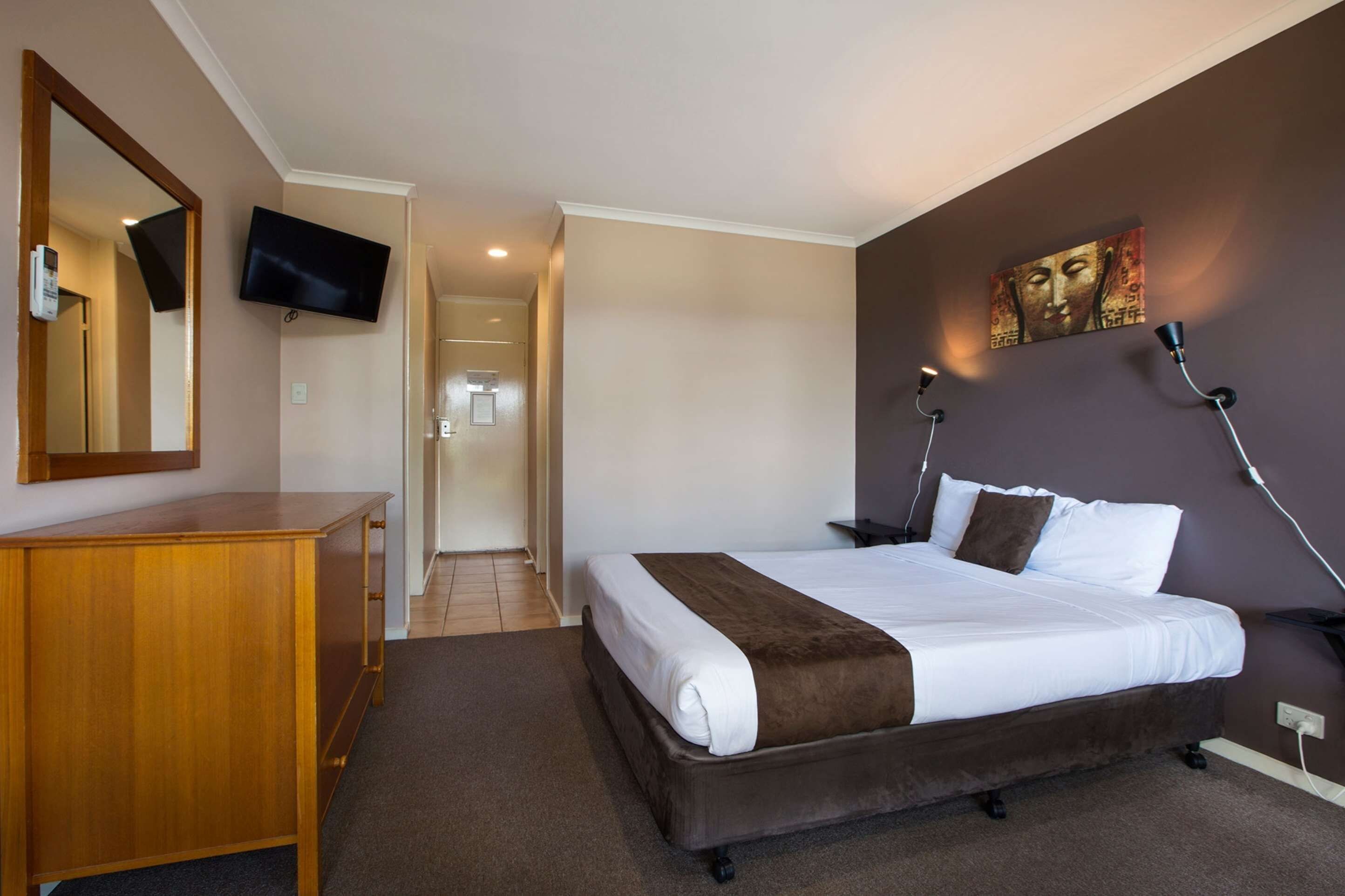 A hotel room with a bed, a wooden dresser, a flat-screen TV, a painting on the wall, and a lamp.