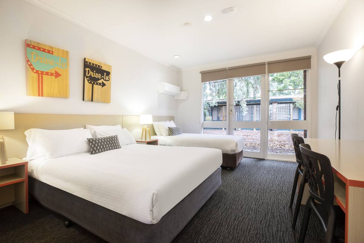 Une chambre d’hôtel avec deux lits, un bureau et une chaise. Elle est équipée de deux climatiseurs fixés au mur et possède une grande fenêtre donnant sur l’extérieur.