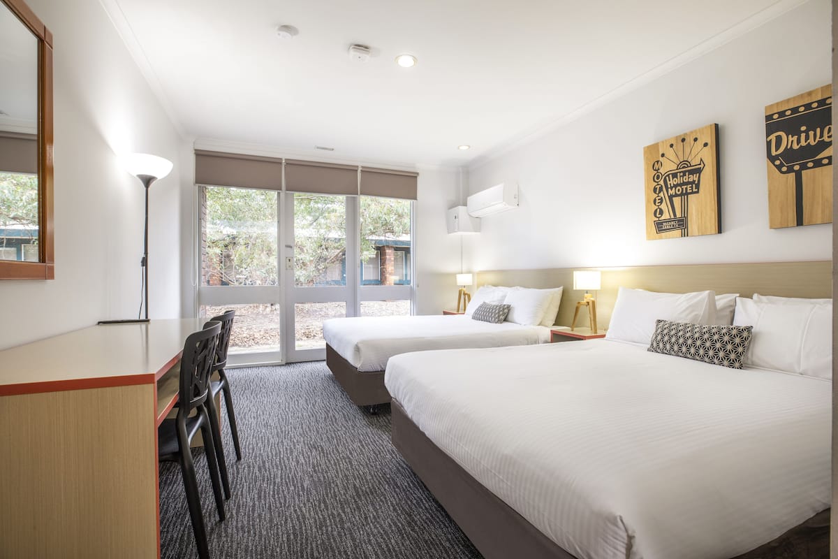 Une chambre d’hôtel avec deux lits, un bureau et une chaise. Il y a deux tables de chevet avec des lampes. La chambre dispose d’une grande fenêtre donnant sur des arbres et un bâtiment situé à l’extérieur.