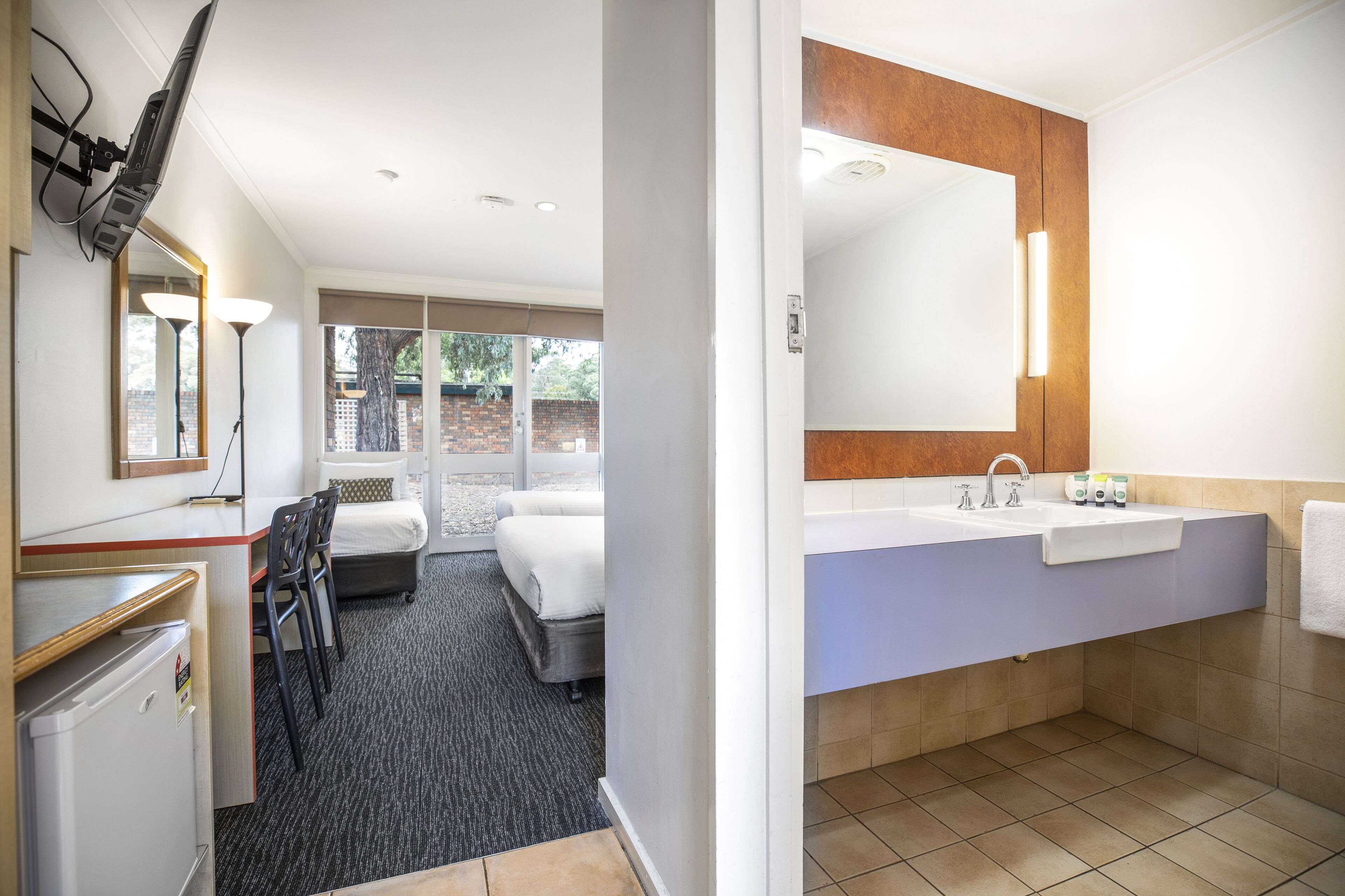 Habitación de hotel con cama, escritorio, silla, refrigerador, espejo grande y baño con lavamanos e inodoro.