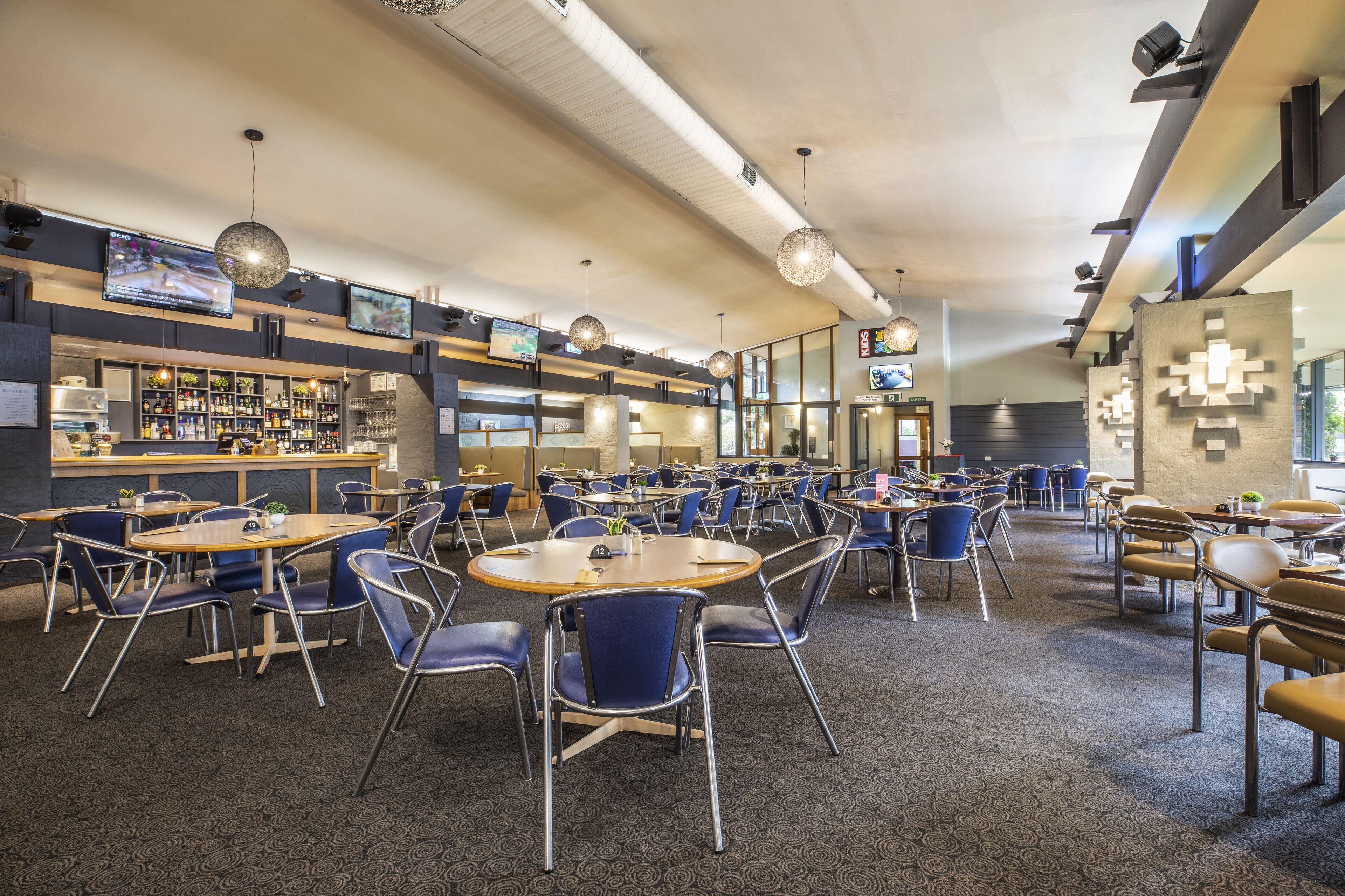 A spacious indoor area with multiple tables and chairs, a bar area with shelves of bottles, and several screens displaying content.