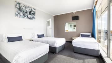 A hotel room with three beds, a large window with a view, a painting on the wall, and a flat-screen TV.
