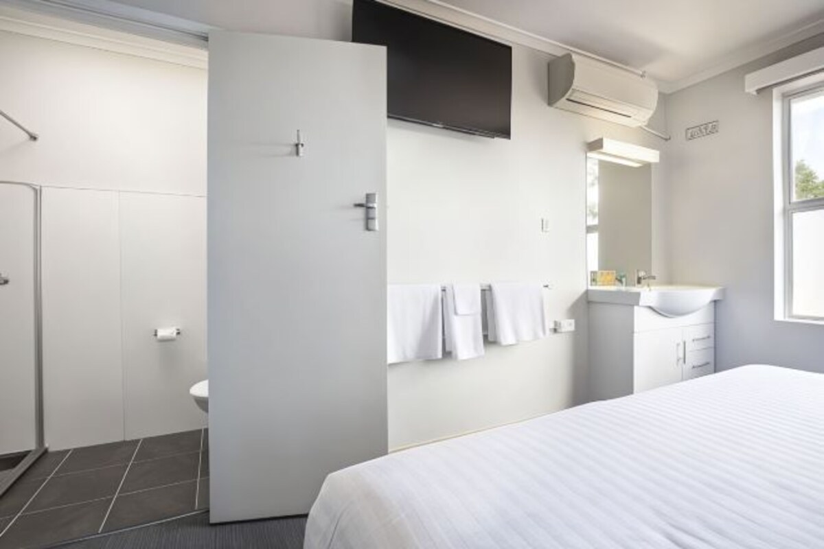 Una habitación de hotel moderna con cama, baño con inodoro y lavamanos, y un ventanal que proporciona luz natural.