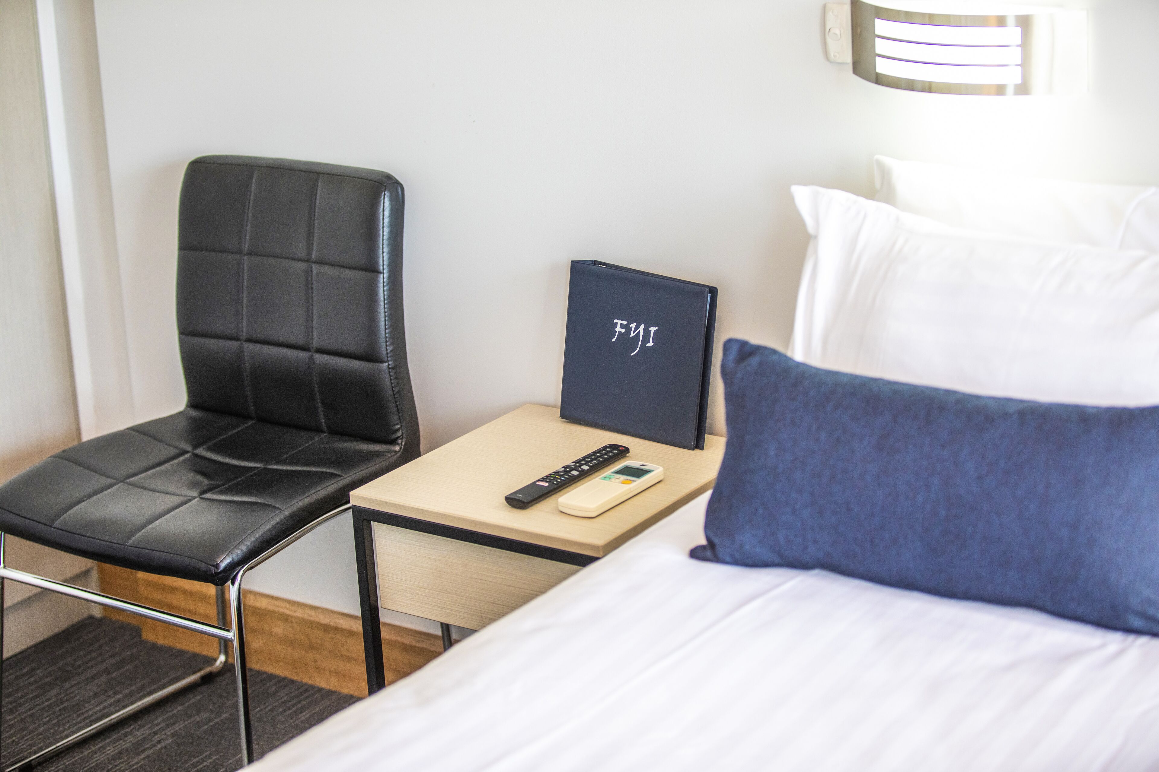 A hotel room with a bed, a black leather chair, a bedside table with a remote control, a book, and a pillow.