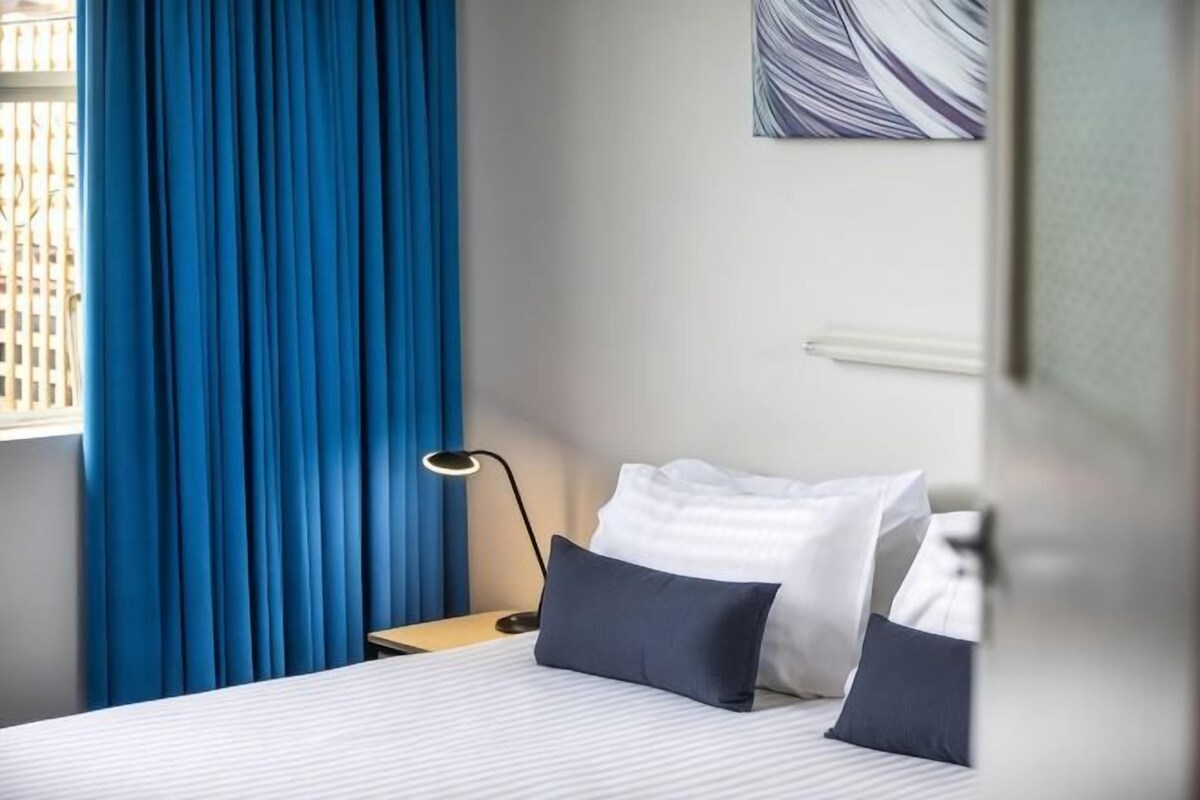Una habitación de hotel con una cama, una mesita de noche, una lámpara y un ventanal con cortinas azules.