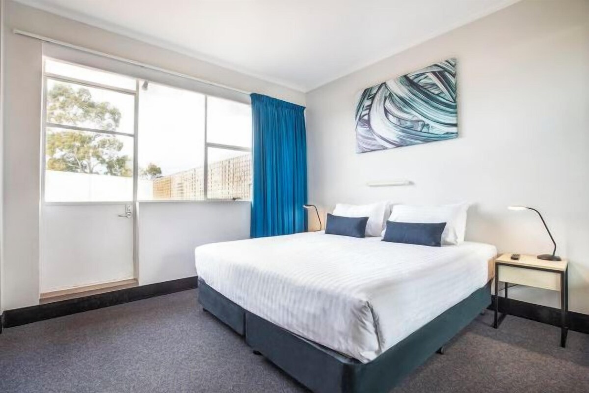 A hotel room with a large bed, a window with curtains, a painting on the wall, and a bedside table with a lamp.