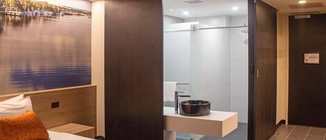 Habitación de hotel moderna con cama, mesita de noche, baño con ducha y un ventanal con vistas al agua.