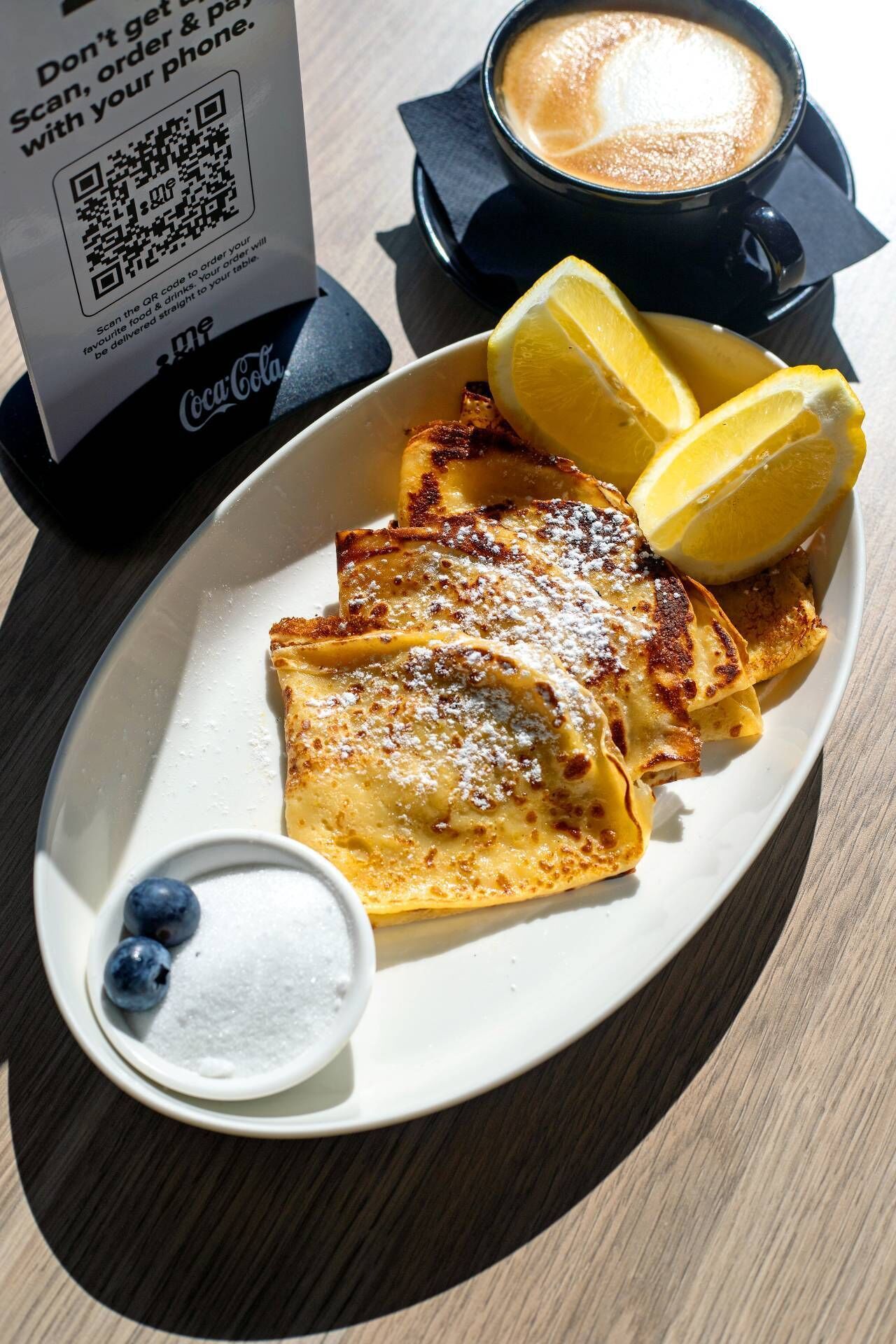 Un plat de crepes amb sucre glas i aranyons blaus, una tassa de cafè i un objecte de Coca-Cola.