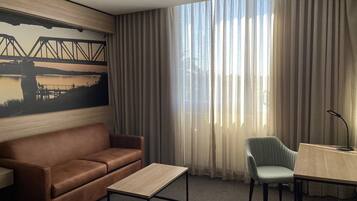 Habitación de hotel con un sofá de cuero marrón, una mesa de centro de madera, un sillón azul claro, un escritorio pequeño con lámpara y un ventanal con cortinas traslúcidas.