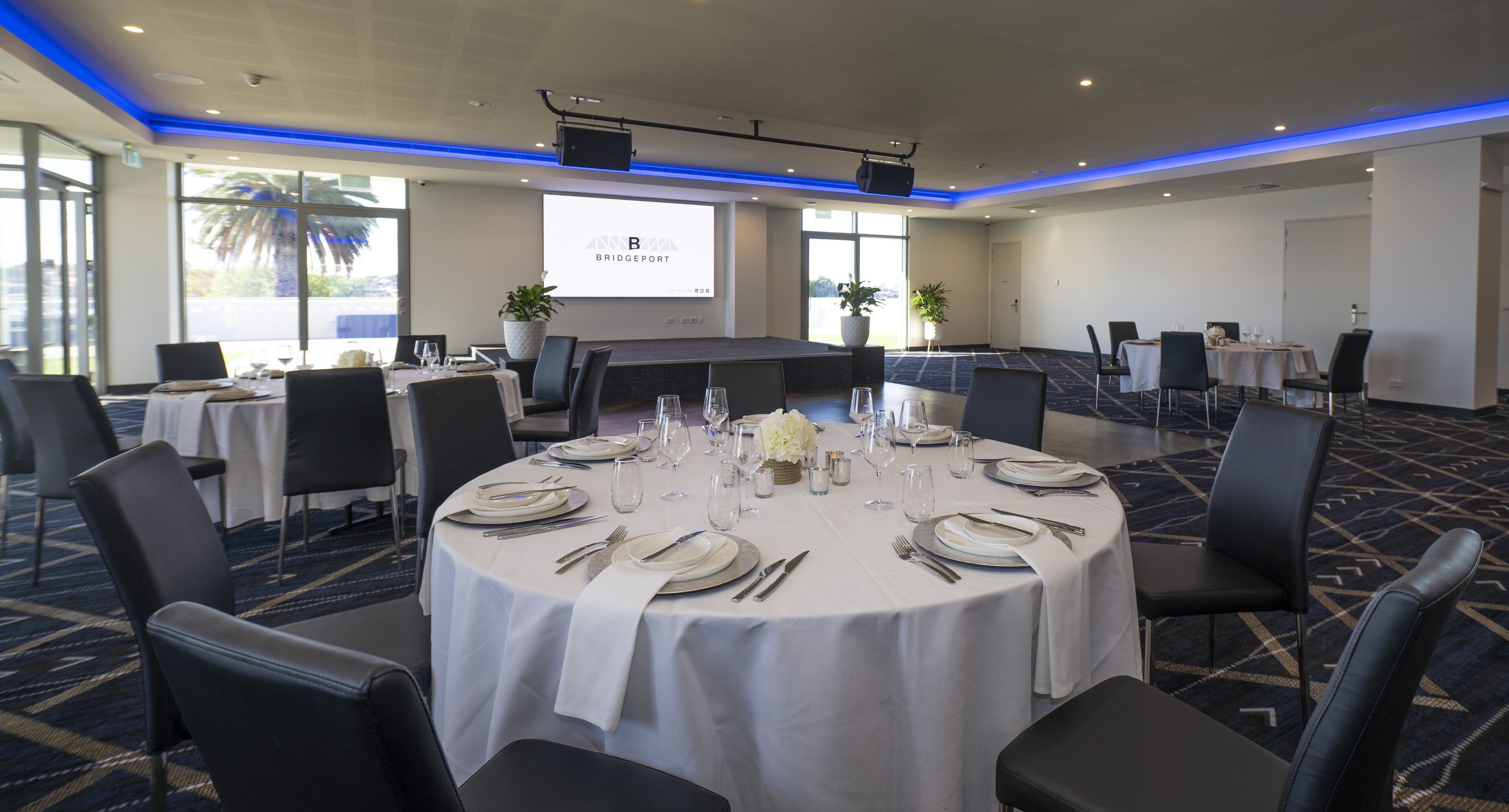A conference room with a large screen, round tables set for dining, and a carpeted floor.