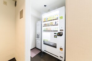 A vending machine with various drinks and snacks, a water dispenser, and a small trash bin in a hallway.
