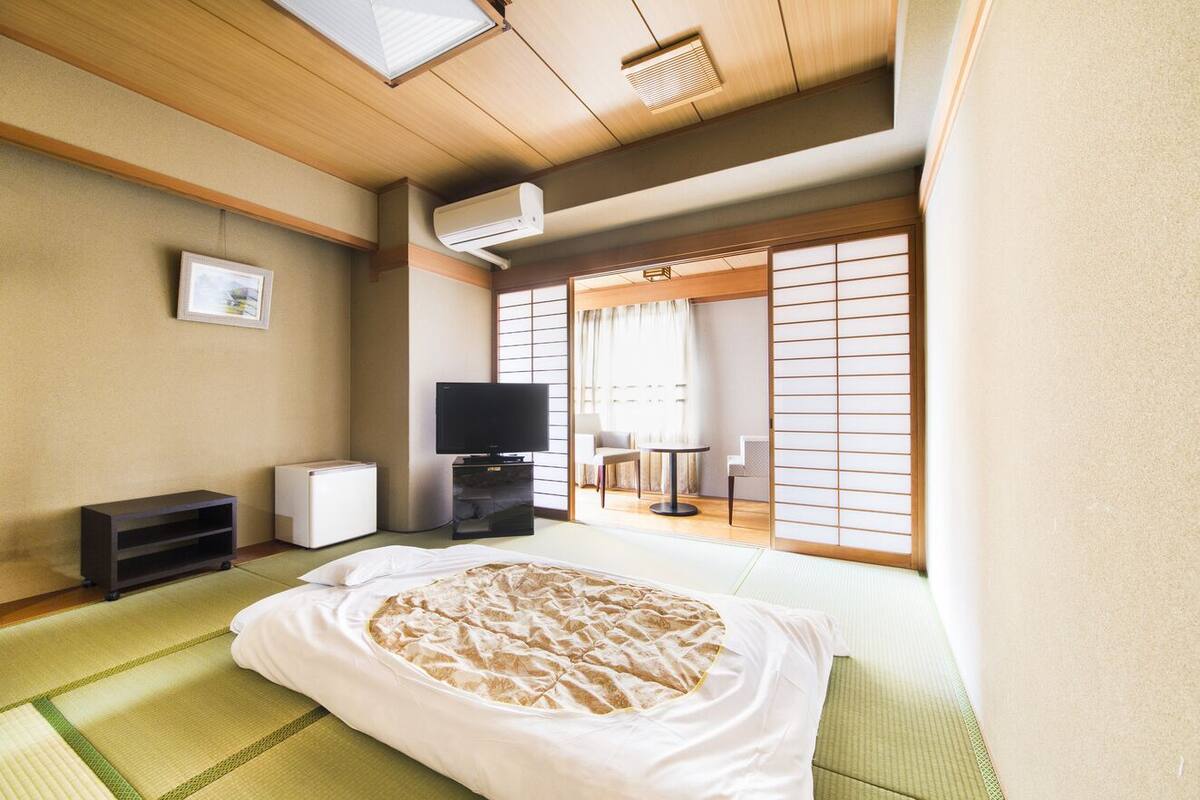 Um quarto japonês tradicional com tatames, uma cama com cobertor estampado, uma televisão, um pequeno armário e portas shoji deslizantes.