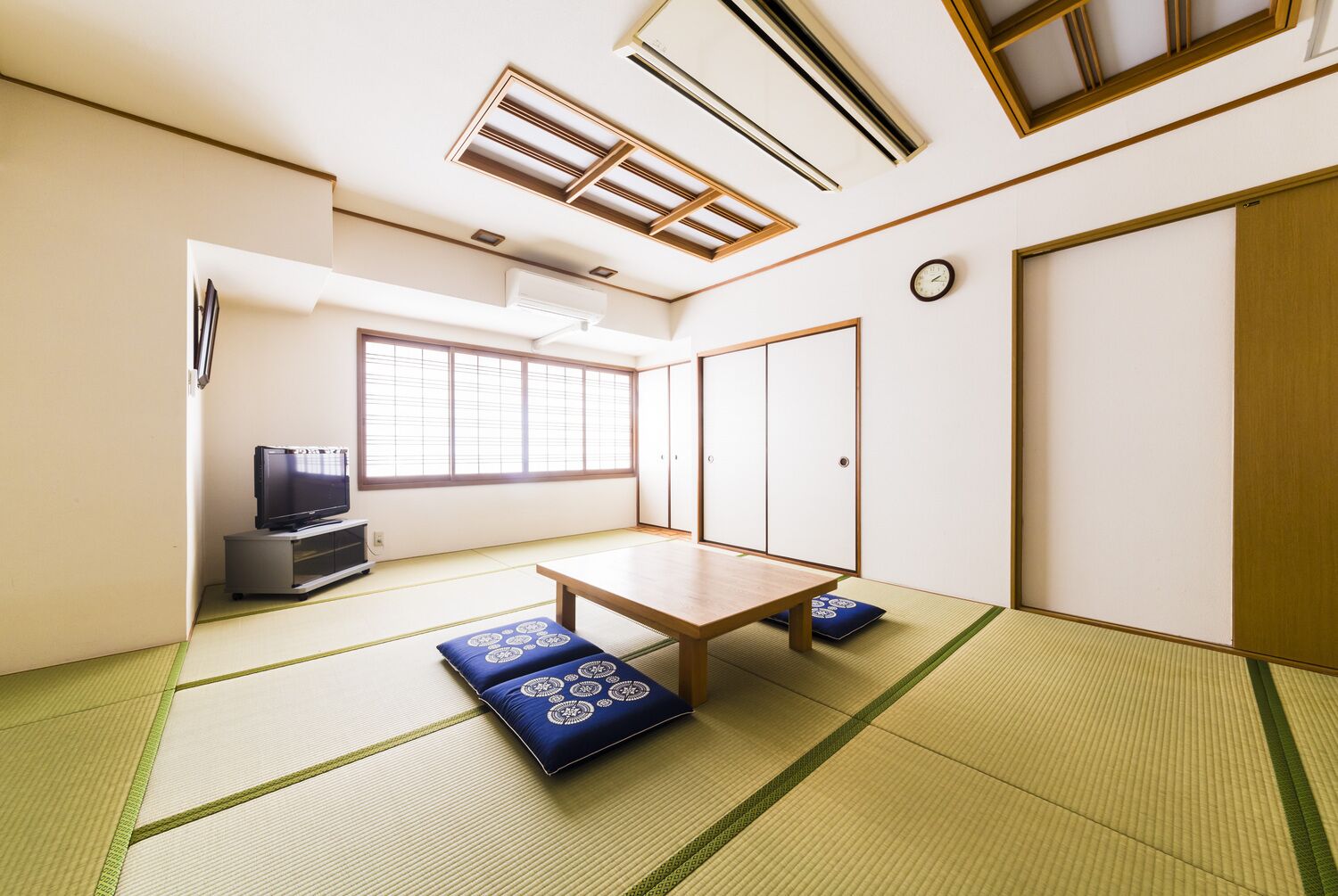 A traditional Japanese room with tatami flooring, a low table, a television, sliding doors, and a window with blinds.