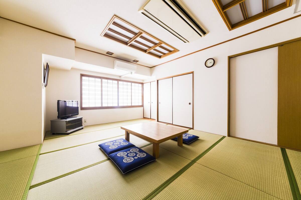 Um quarto japonês tradicional com tatames, uma mesa baixa, televisão, portas de correr e uma janela com persianas.