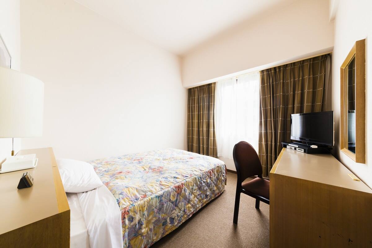 Quarto de hotel com cama, escrivaninha com luminária, cadeira, televisão e janela com cortinas.
