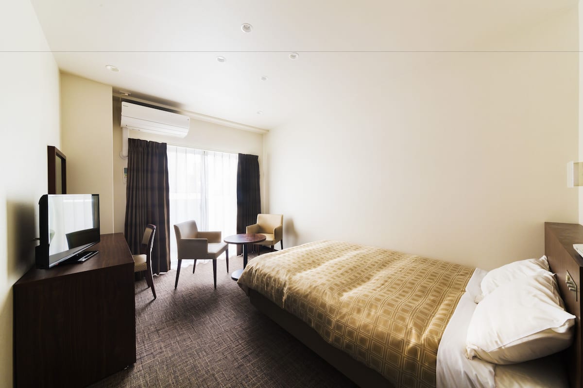 Quarto de hotel com uma cama, televisão em móvel, escrivaninha e duas cadeiras.