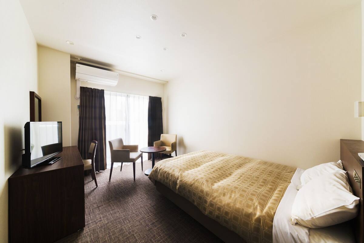 Quarto de hotel com cama, televisão, escrivaninha, cadeira e janela com cortinas.