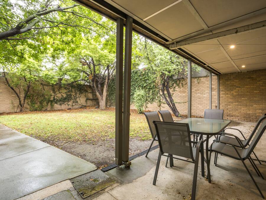 An outdoor patio with a glass table and chairs, a brick wall, and a grassy area with trees.