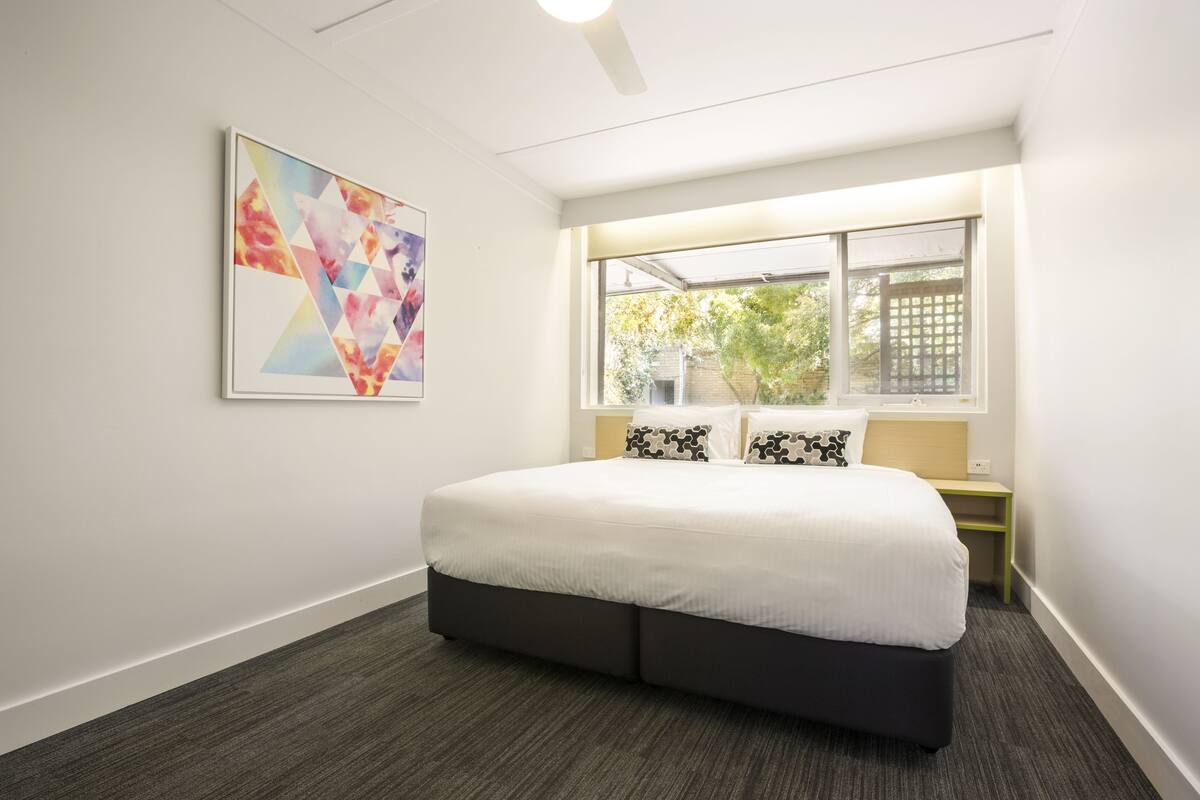 A modern hotel room with a large bed, a window with a view of greenery, and a colorful abstract painting on the wall.