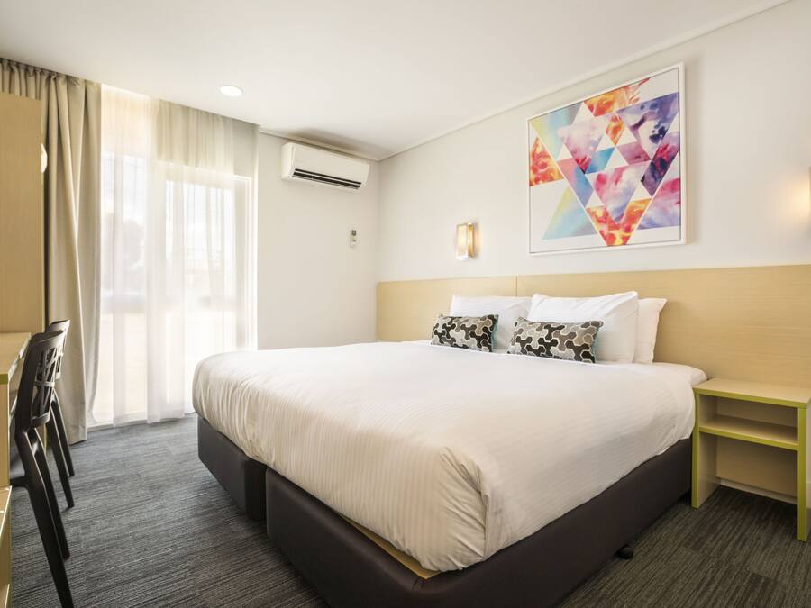 A hotel room with a large bed, a desk, and a chair. There is a window with curtains, an air conditioning unit, and a wall-mounted light fixture.