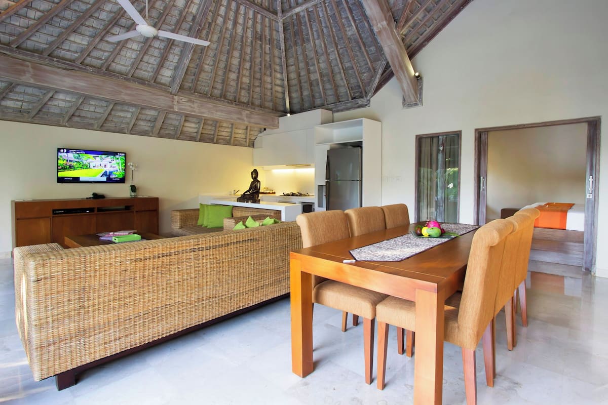 A spacious living area with a high ceiling, a dining table with chairs, a sofa, a kitchen area, and a television.
