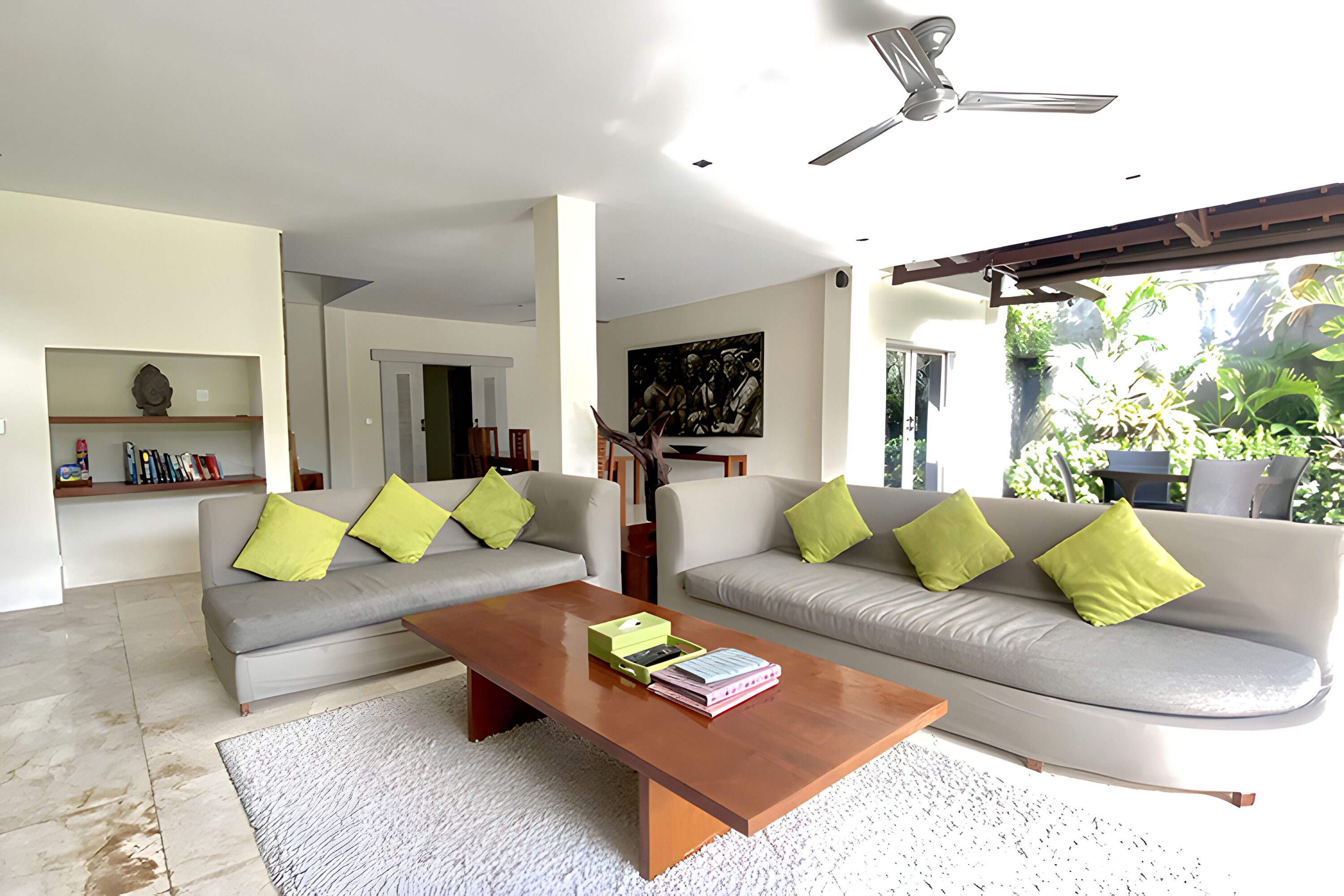 A modern living room with two grey sofas, each with a green pillow, a wooden coffee table, and a white rug. There is a ceiling fan and a painting on the wall.