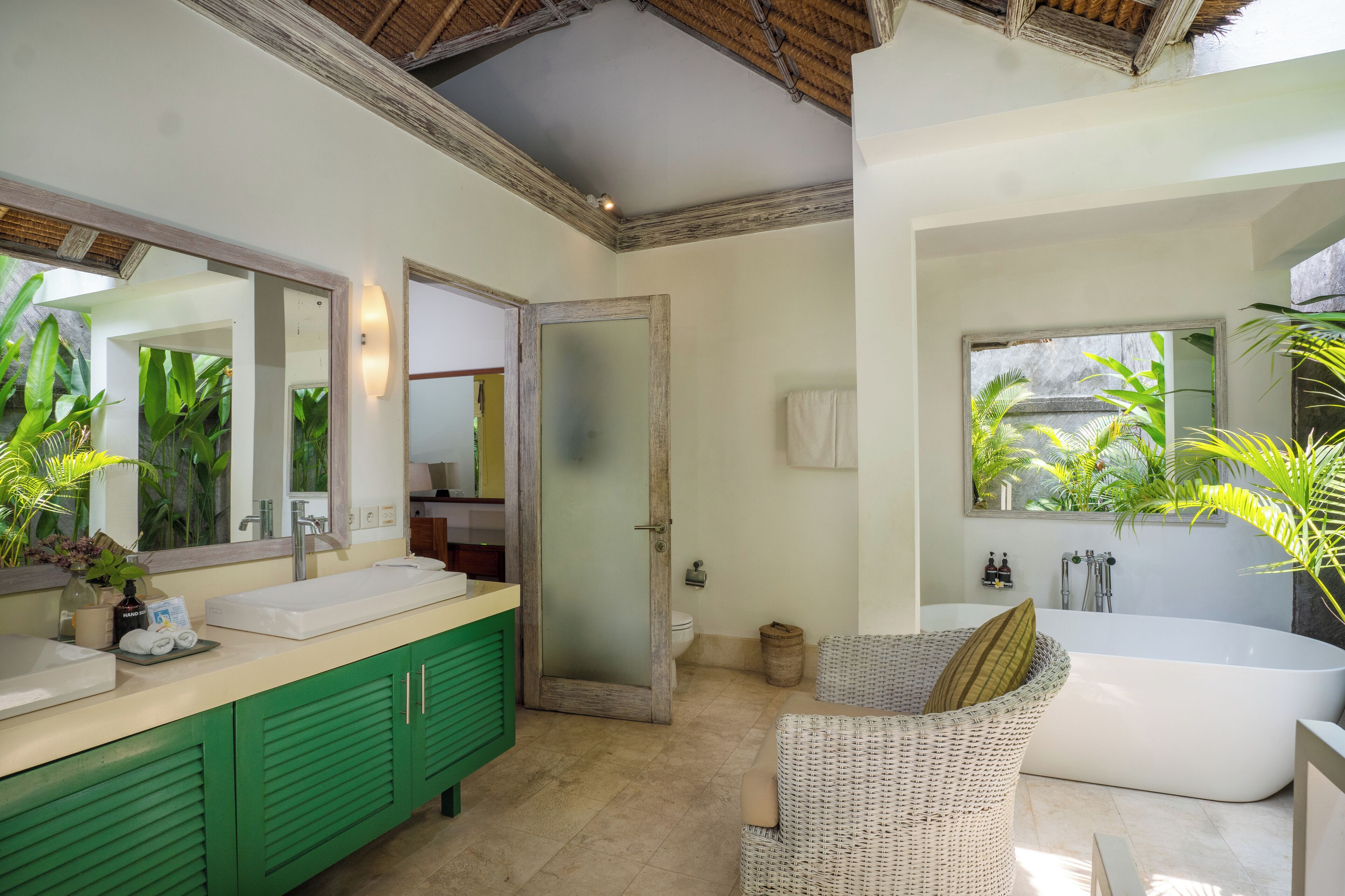 Un baño con un espejo grande, gabinetes verdes, una bañera blanca y una silla de mimbre.