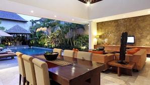 Uma sala de jantar moderna com mesa e cadeiras de madeira, piscina e área de estar com sofás e televisão.
