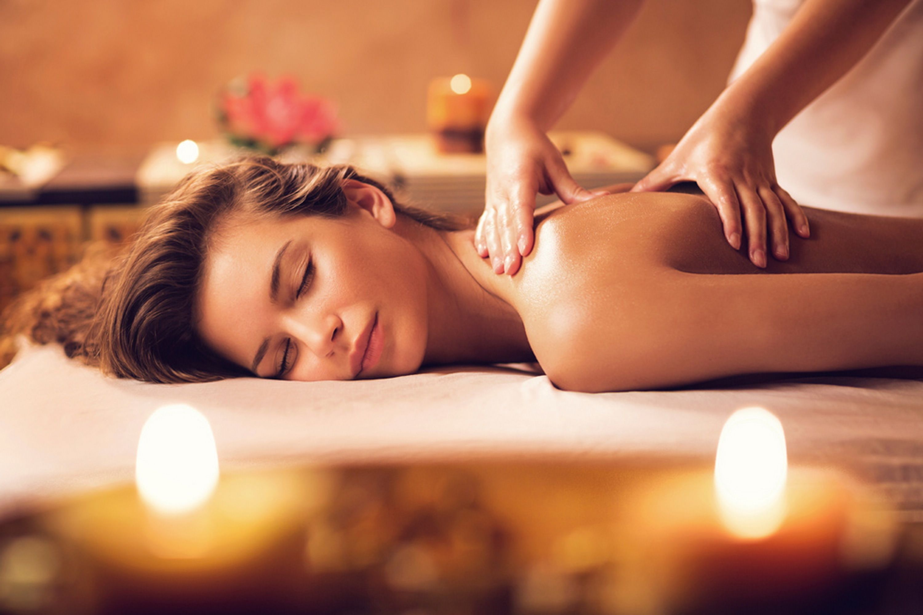 Eine Person erhält eine Massage auf einem Bett, im Hintergrund sind Kerzen und Blumen zu sehen.