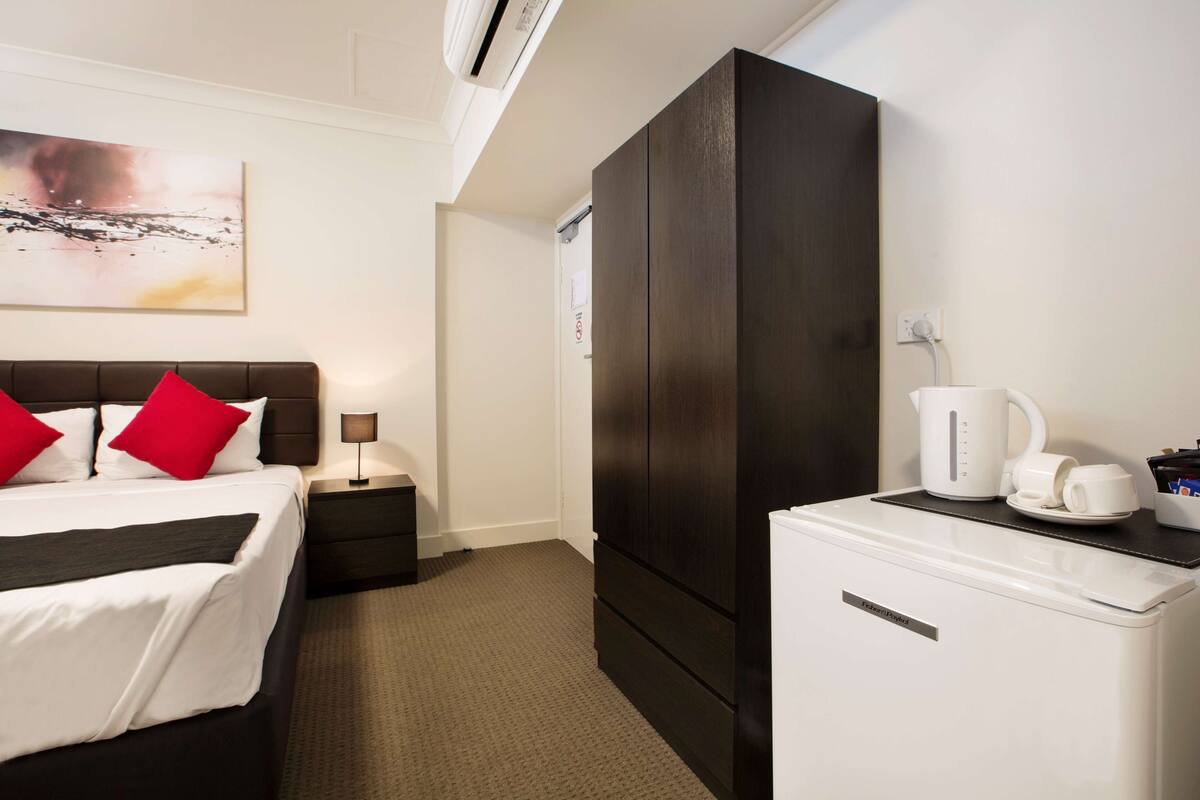 A hotel room with a bed, a nightstand, a wardrobe, a coffee maker, and a small refrigerator.