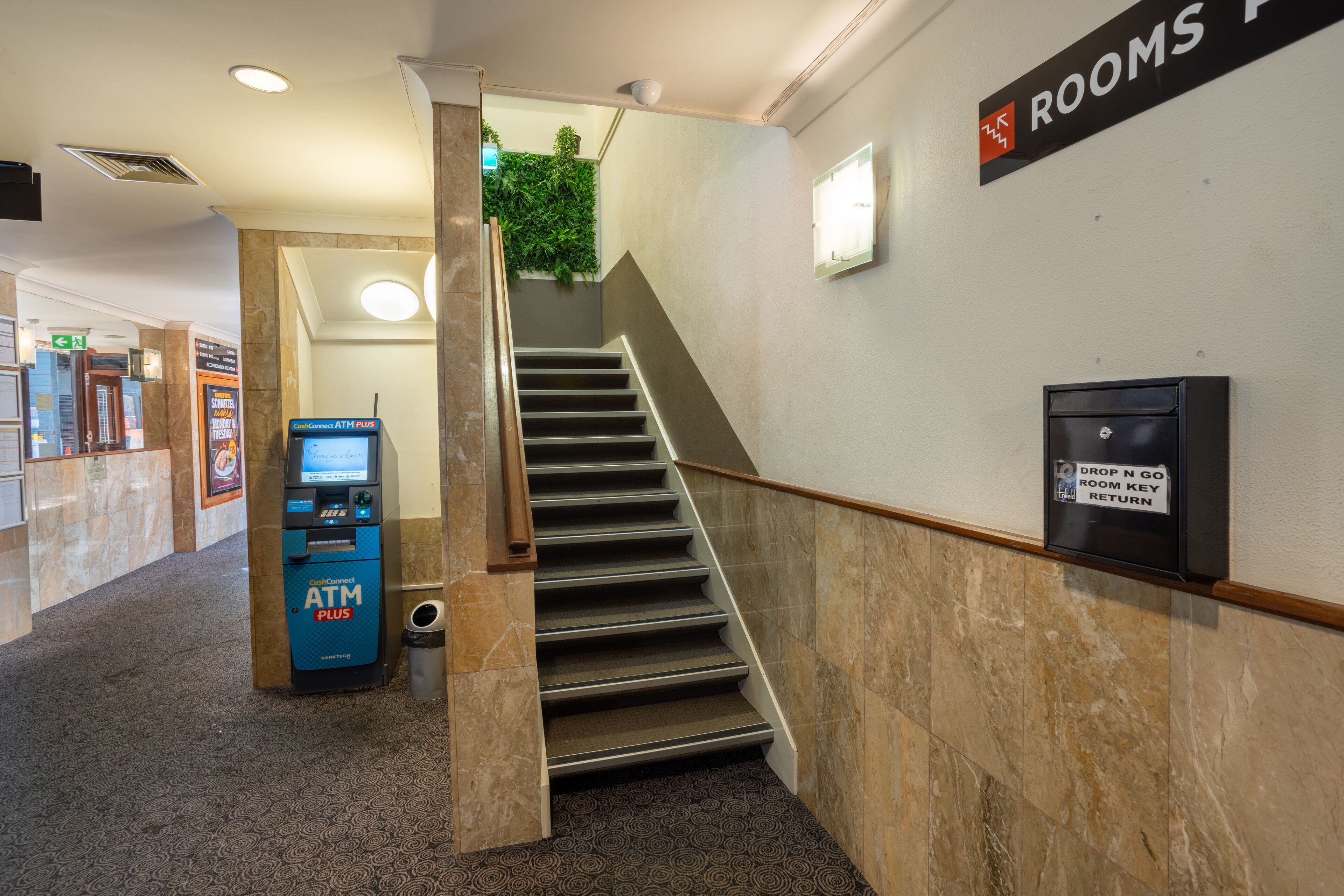 Een hotellobby met een trap naar de kamers, een geldautomaat en een bord dat de locatie van de sleuteluitgifte aangeeft.