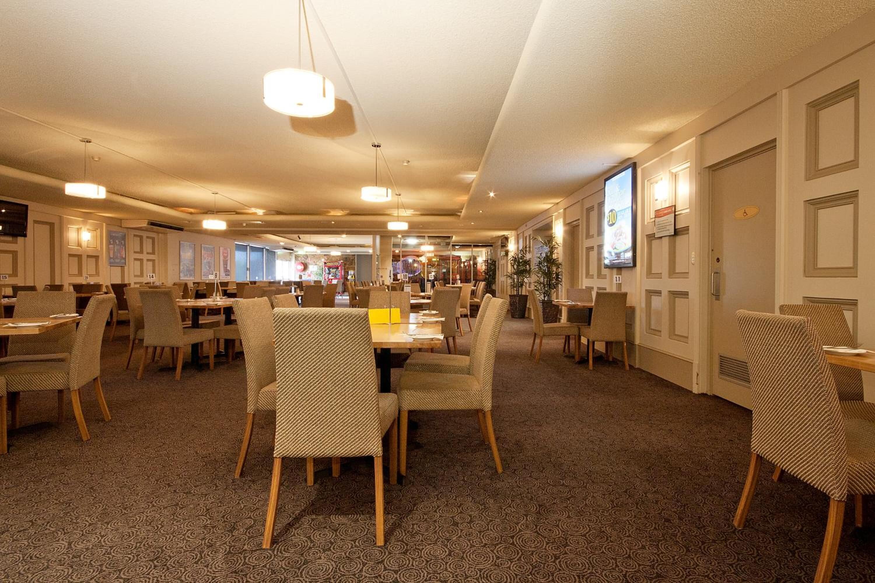 A hotel lobby with multiple dining tables and chairs, a television screen, and a patterned carpet.