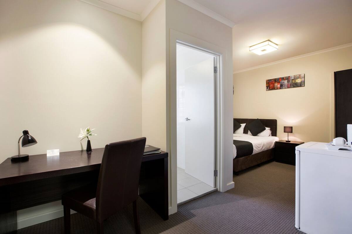 A hotel room with a desk, chair, bed, bedside table, lamp, and a refrigerator.