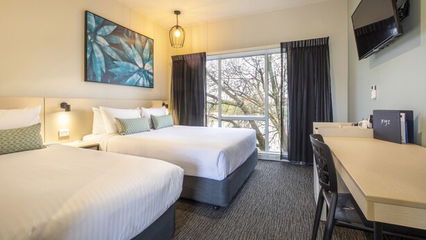 A hotel room with two beds, a desk, a TV, and a large window with curtains. - Nightcap at Excelsior Hotel (Thomastown)