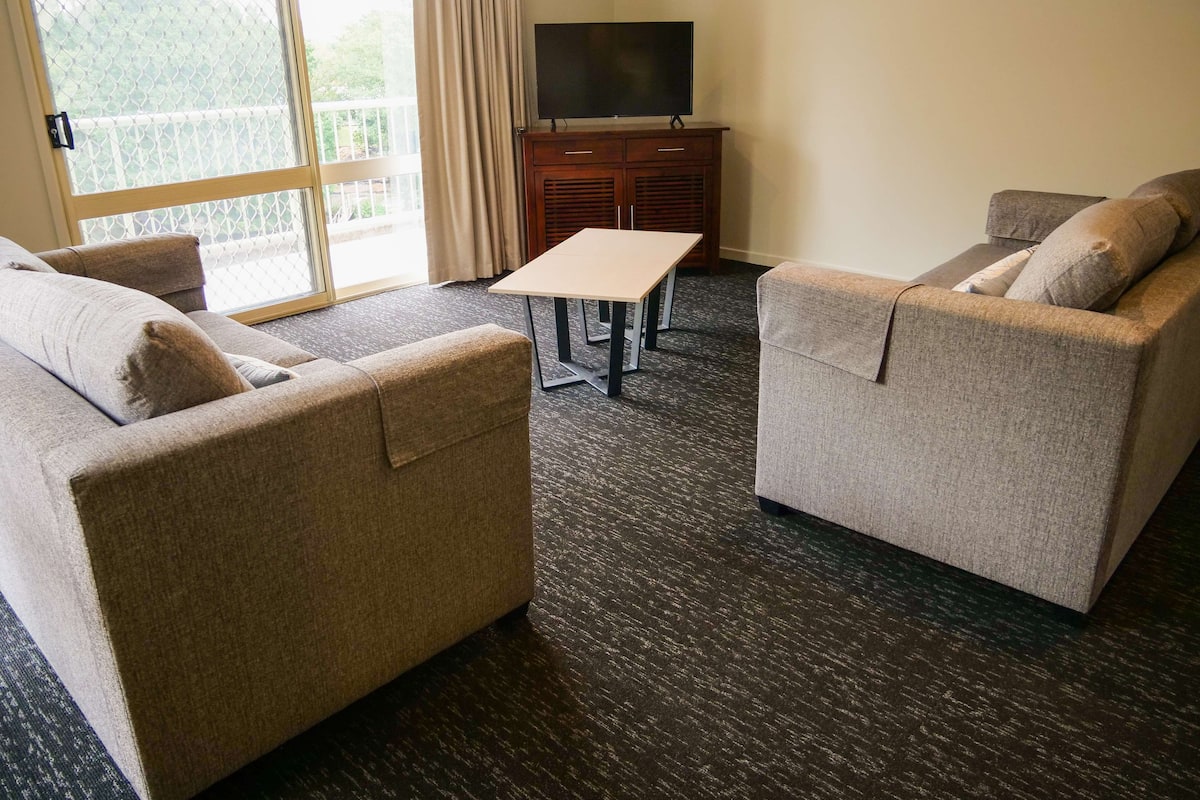 Habitación de hotel con dos sofás, una mesita, un televisor en un mueble bajo y un ventanal con cortinas.