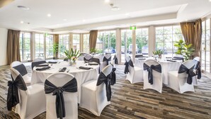 Un salón de conferencias con mesas redondas, sillas blancas con cintas negras, ventanas amplias y suelo alfombrado.