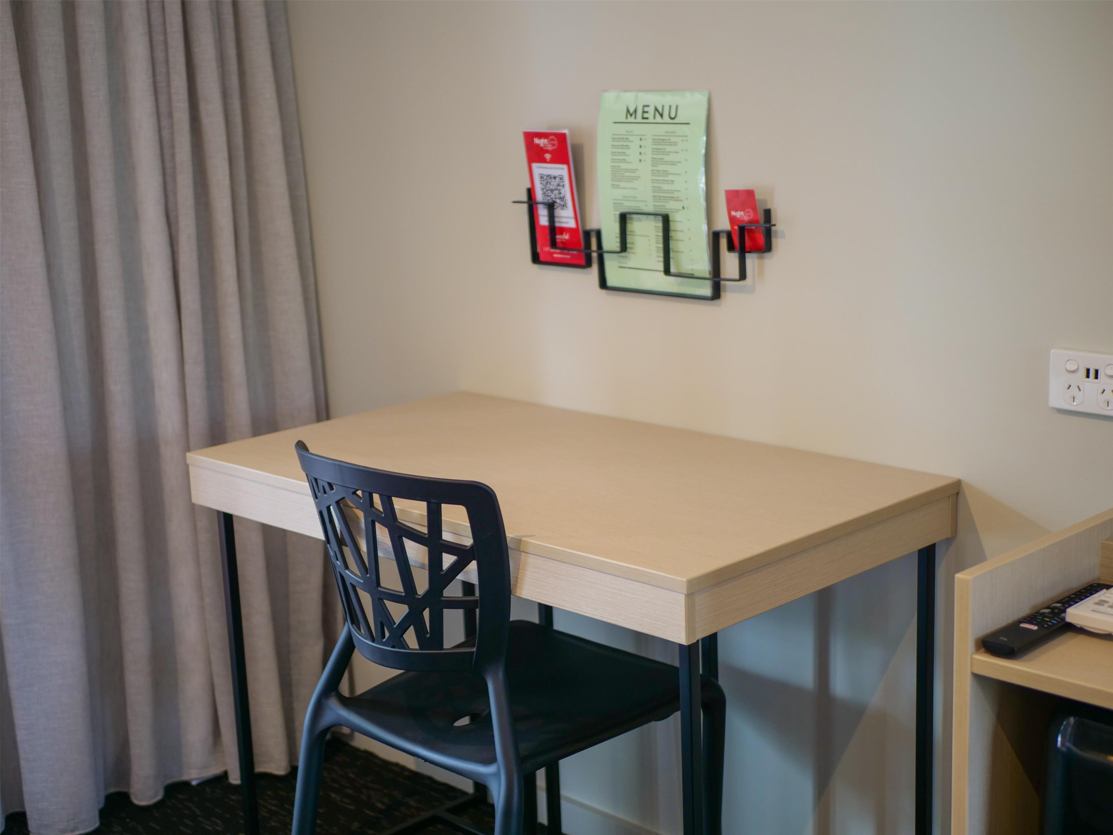 A hotel room with a single bed, a desk with a chair, a wall-mounted menu, and a fire extinguisher.