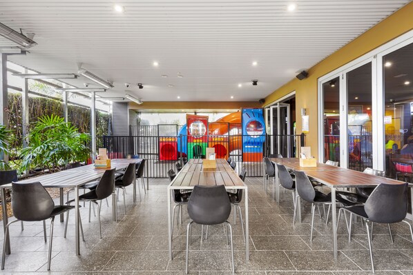 An outdoor dining area with a long wooden table and black chairs, surrounded by a fence with a playground visible in the background.